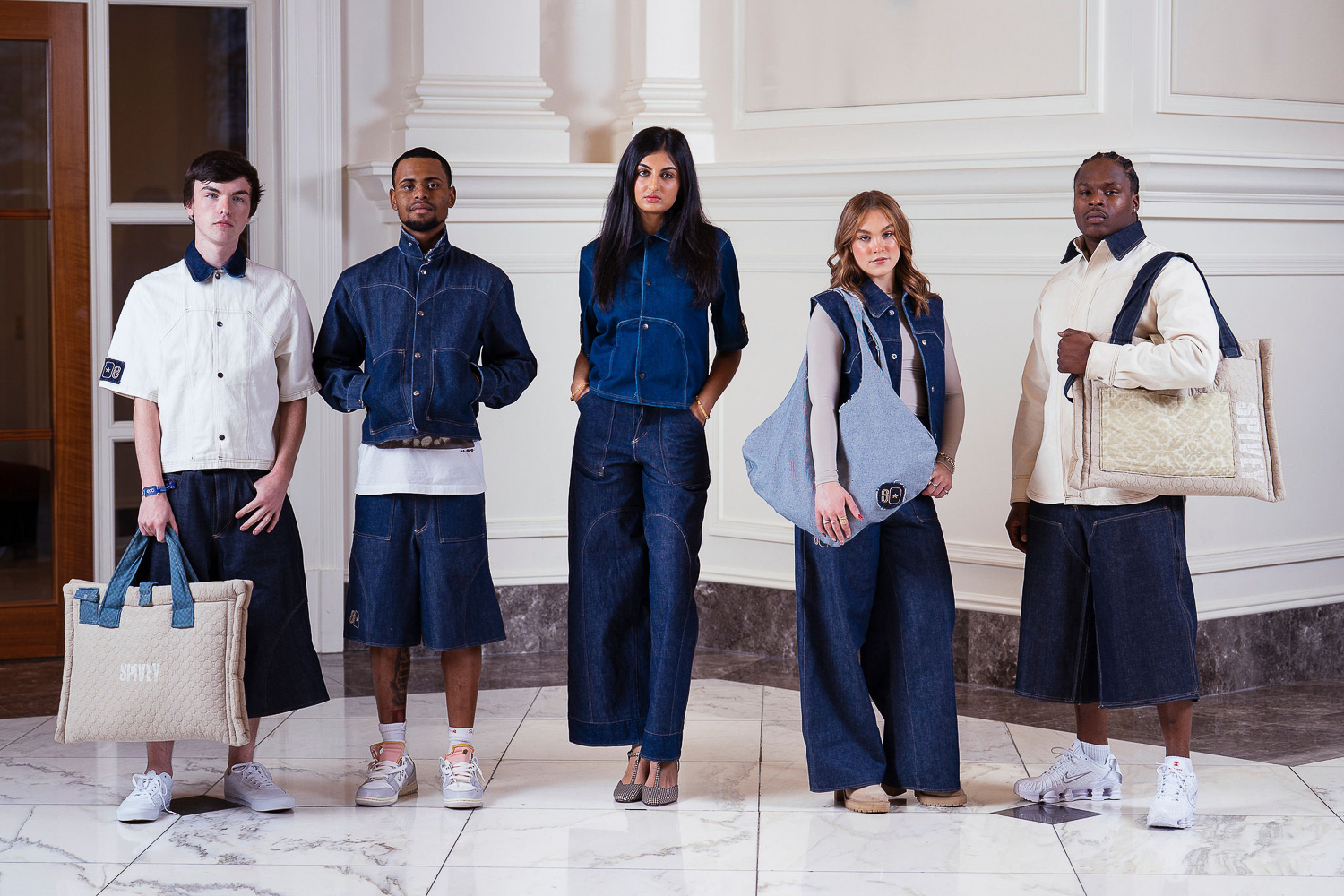 Five people stand indoors against white paneled walls, modeling matching blue denim outfits and holding large designer bags. The group includes both men and women, all facing forward on a marble floor.