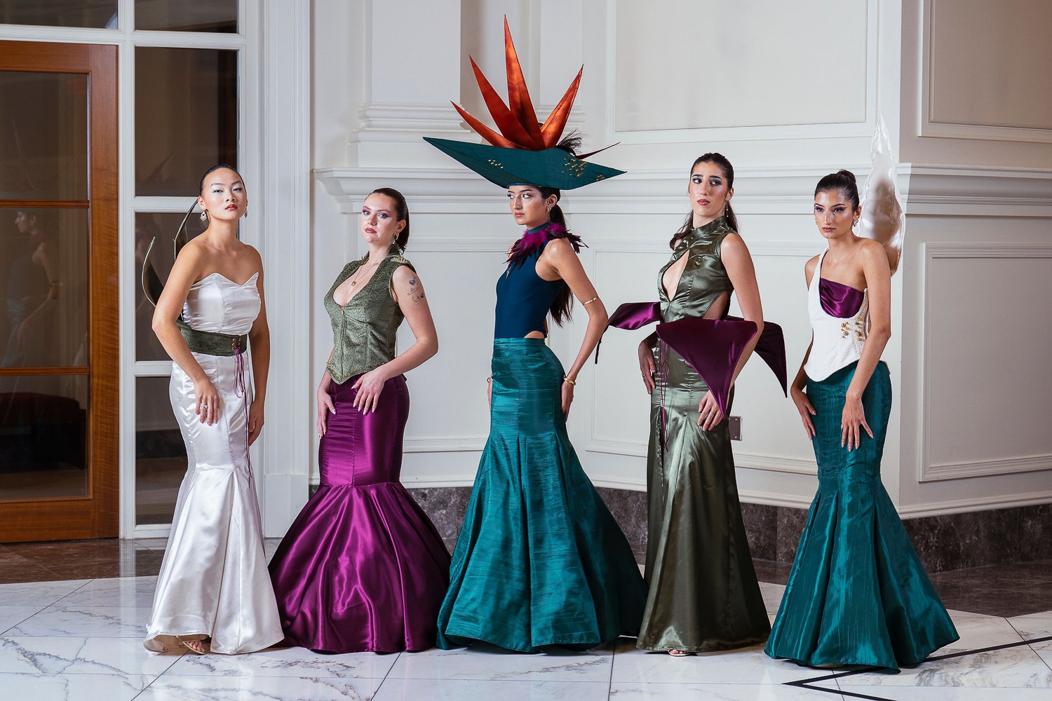 Five models pose in elegant, sculptural gowns in jewel tones. One wears a dramatic hat with orange spikes. The setting is a bright, modern interior with white walls, marble floors and large windows.
