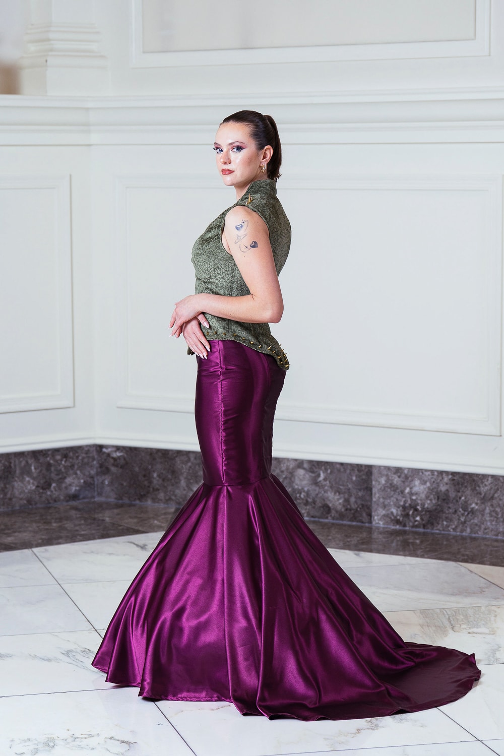 Rachel Kelly poses indoors, wearing a fitted, floor-length, purple mermaid skirt and an olive green textured top. She stands on white marble flooring against white paneled walls with a marble baseboard.