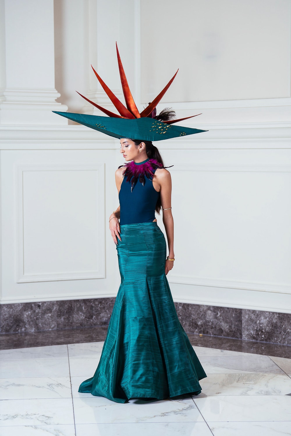 A model wears a dramatic teal gown and a large, sculptural hat with red-orange spikes and green accents, resembling a tropical plant. They stand in an elegant, white-walled interior with marble flooring.