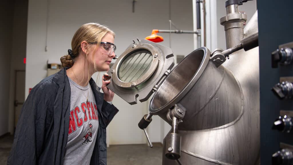 Lindsay Davis stands wearing safety glasses holding open a small circular door on a piece of machinery and peering inside.