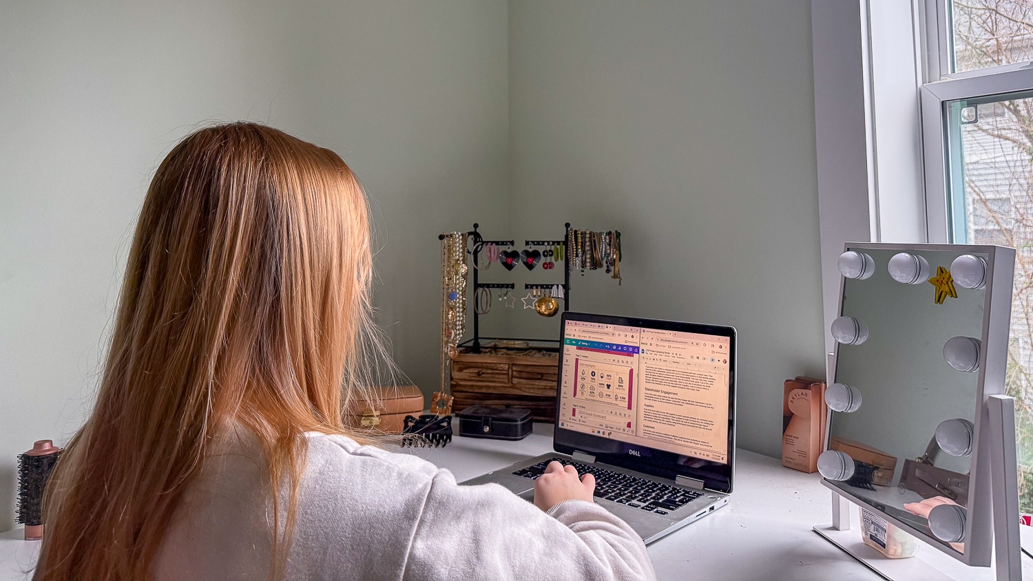Nikki Fiske sits at a white desk using a laptop. The image looks over her shoulder to show the back of her head, her shoulders, and a laptop screen which has a presentation in progress on it. The desk has jewelry, a makeup mirror with lights, and a window showing trees outside.