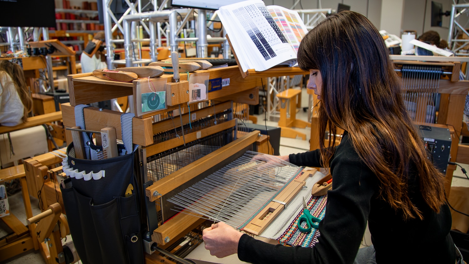 Edie Youngblood sits at a wooden loom, weaving with a shuttle in the Wilson College of Textiles weaving studio. Open books, color charts and weaving tools surround her.