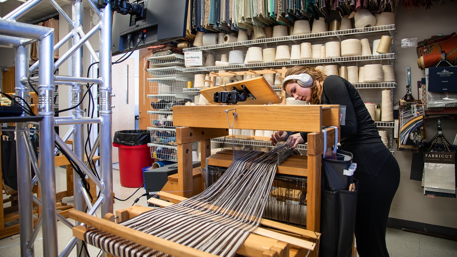 A student in a Wilson College of Textiles studio wears headphones while threading a wooden loom to prepare it for weaving; shelves behind hold yarn and fabric samples.