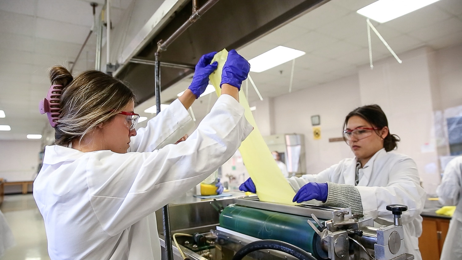 Two people in lab coats, goggles and purple gloves are working together in a laboratory, feeding a dyed yellow textile sample through a rolling machine in a Wilson College of Textiles lab.