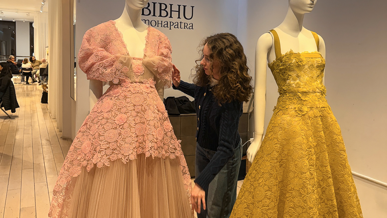 Ashley Rose Phillips adjusts a pink floral gown displayed on a mannequin. To the right is another mannequin with a yellow floral dress.