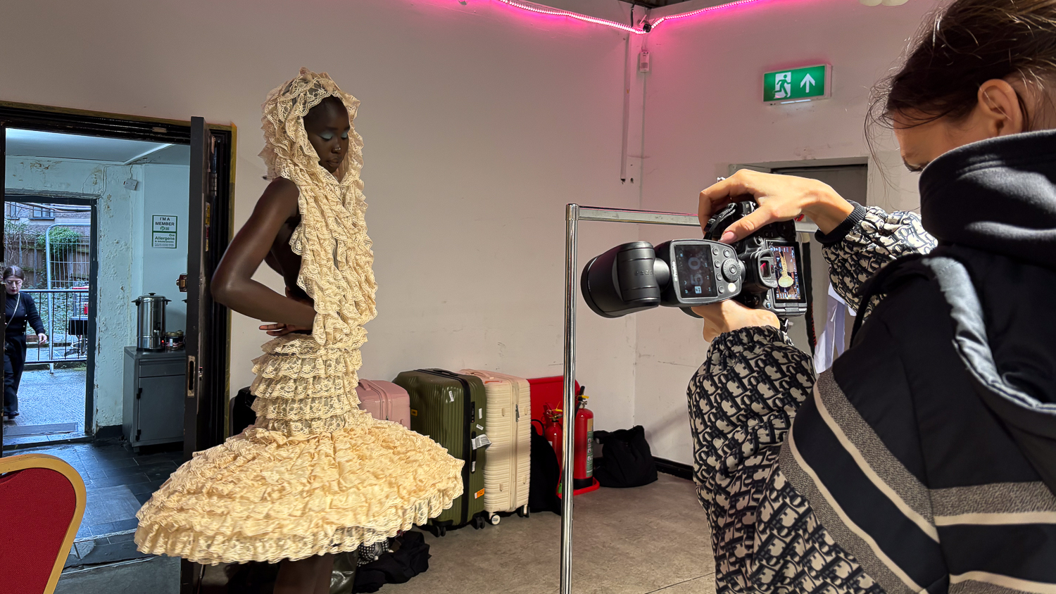 Grace Chen takes a photo of a model in a backstage area. Chen is on the far left of the frame with a large professional camera, and the model poses a few feet away in a frilled white dress.