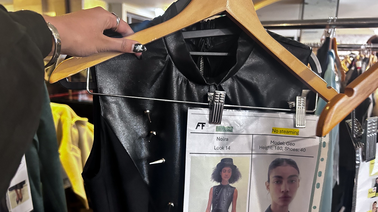 A hand holds a hanger with a black garment and paper attached with a model's information and photo.