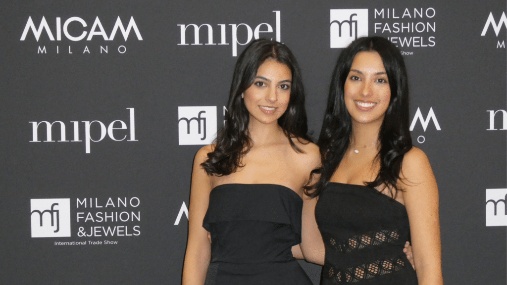Two students stand in front of a display for Milan Fashion Week. They are both wearing black tube tops and have medium-length black hair, smiling at the camera with their hands around each others backs.