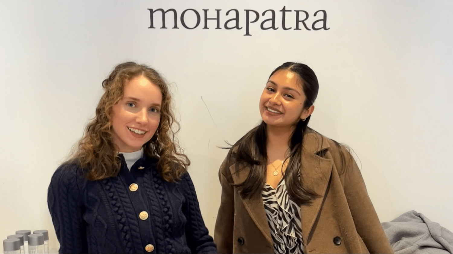 Two students stand in front of a white wall with black text on it. They both smile at the camera in the middle of the frame.