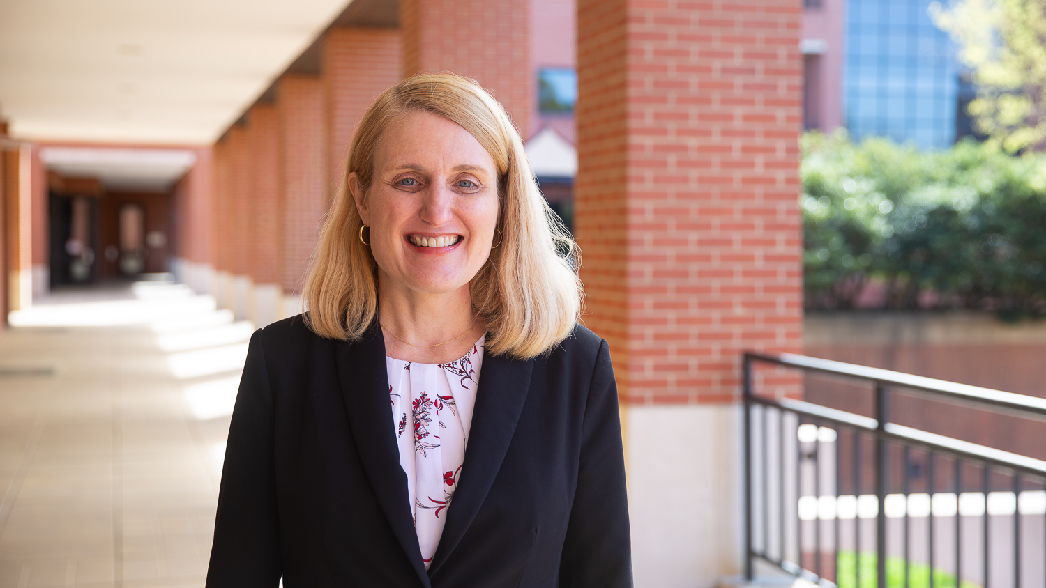 Kristin-Thoney Barletta smiles and looks at the camera on a sunny day. She stands in a breezeway and some of the Wilson College of Textiles main building is visible in the background.