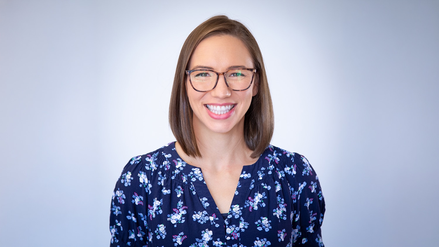A headshot of Wilson staff Bridget Inzirillo. She has medium length brown hair, a brown framed glasses, a blue flowery blouse, and a big smile.
