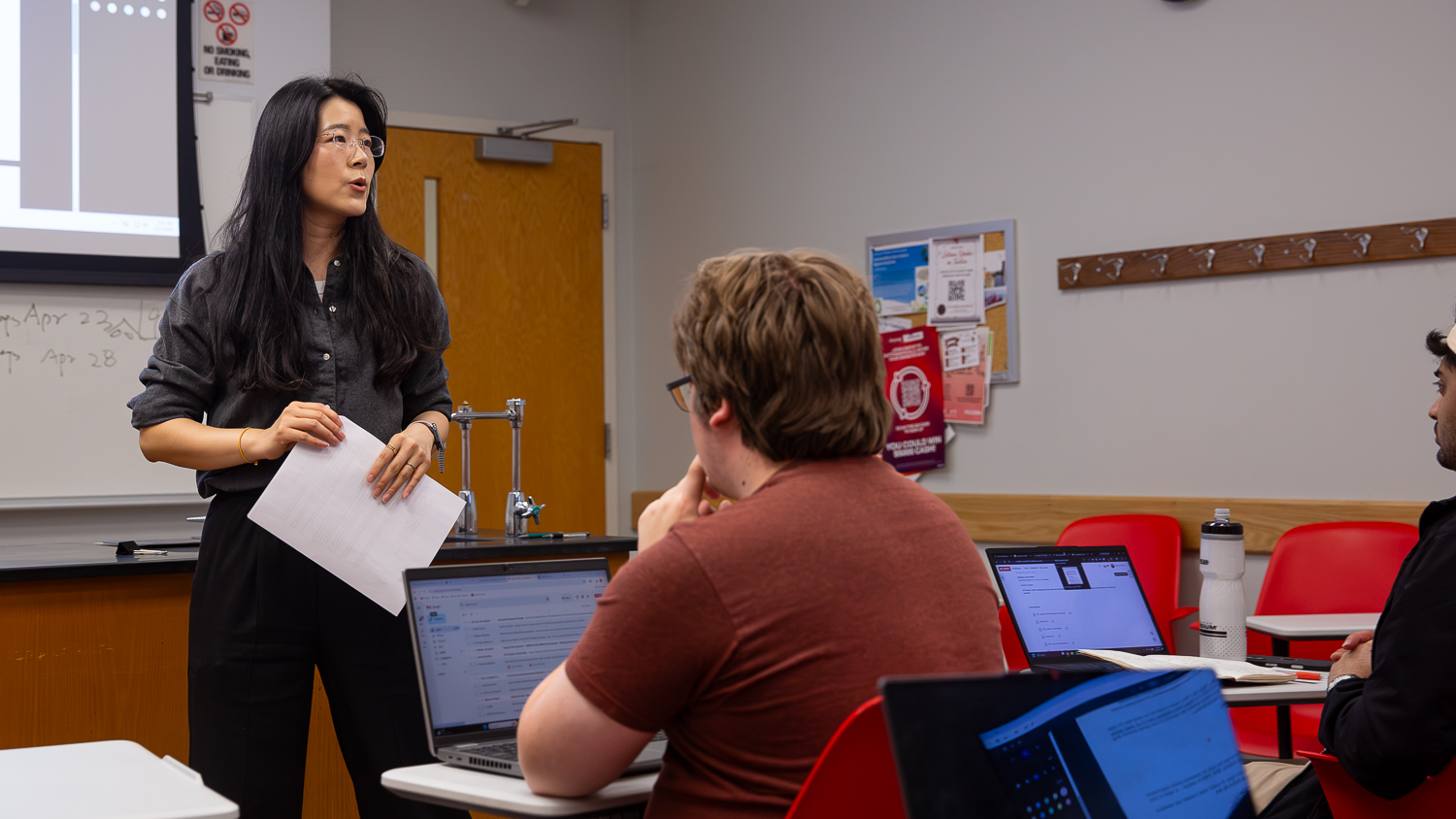 Mengmeng Zhu stands at the front of a classroom holding papers and speaking, while students with laptops sit at desks and listen. The classroom features red chairs, a whiteboard and a bulletin board.