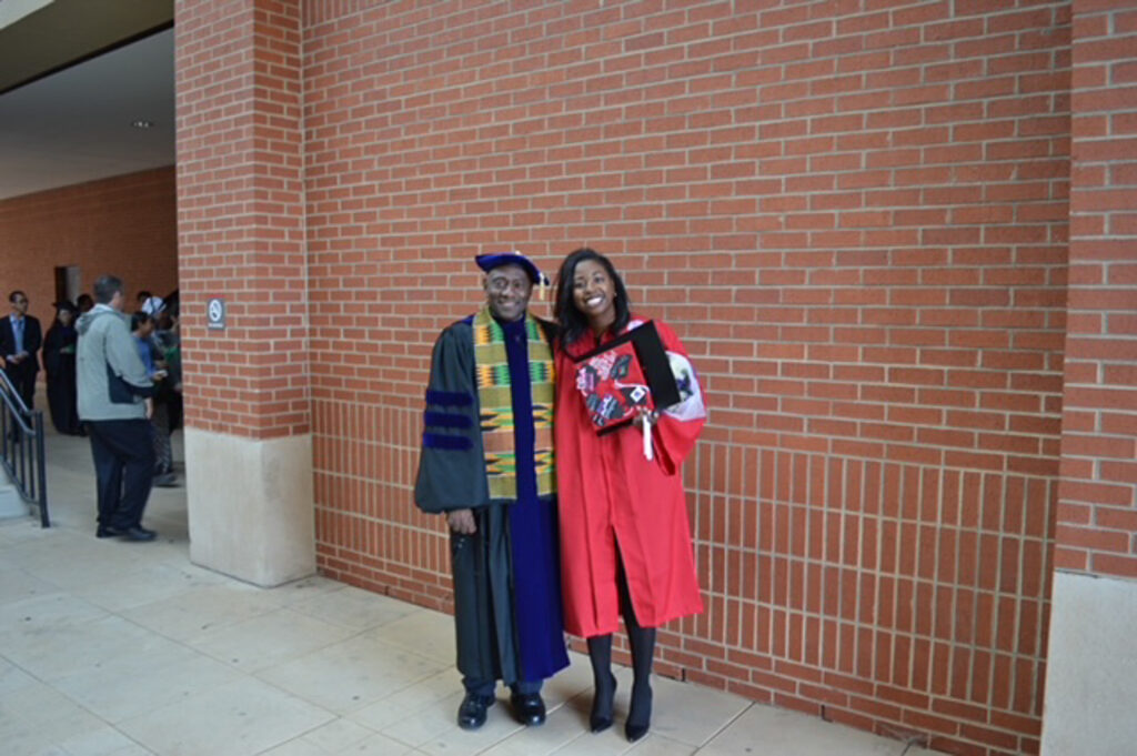 Courtney Priester-Funderburk (right) stands next to Harold Freeman in front of a red brick wall. Both are wearing commencement regalia and smiling a the camera.