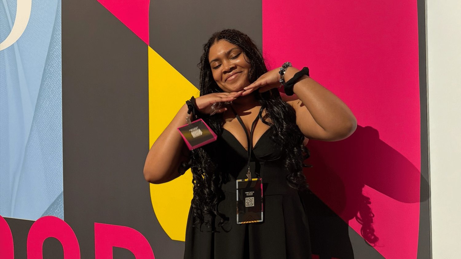 Celia Fennell wears a black dress and stands smiling with her hands under her chin in front of a colorful, geometric wall with large yellow, pink, and black shapes and bold text. She is wearing event badges around her neck.