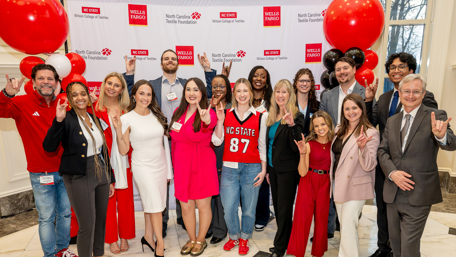 A group of people pose smiling in front of a step-and-repeat banner with NC State Wilson College of Textiles and Wells Fargo logos. Some have their "wolves up." Red and white balloons decorate the scene.