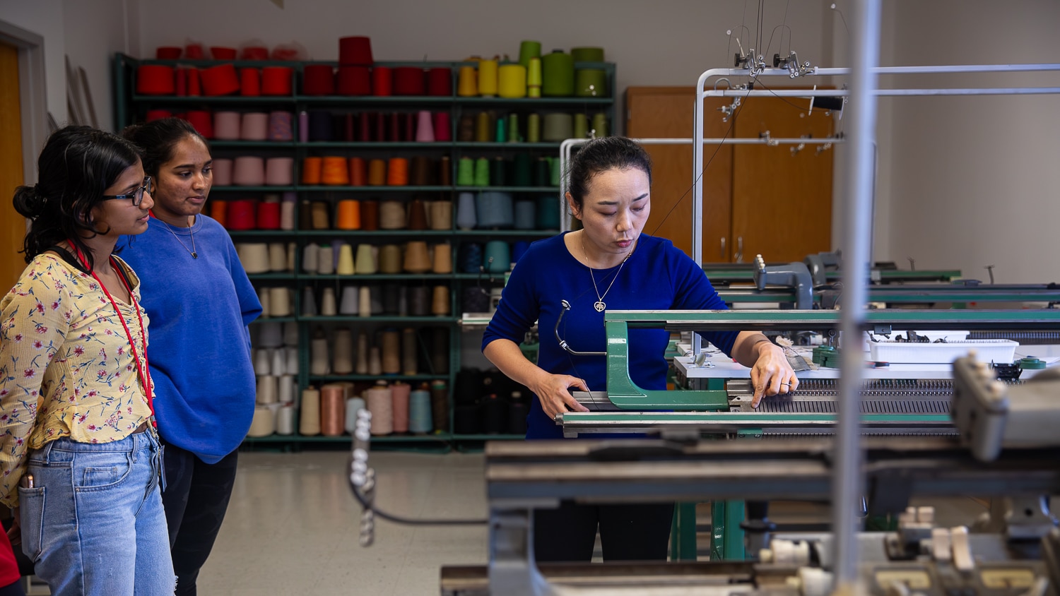 Xiaomeng Fang demonstrates how to use a knitting machine to two students in a knitting lab at the Wilson College of Textiles. Shelves with colorful spools of thread are visible in the background.