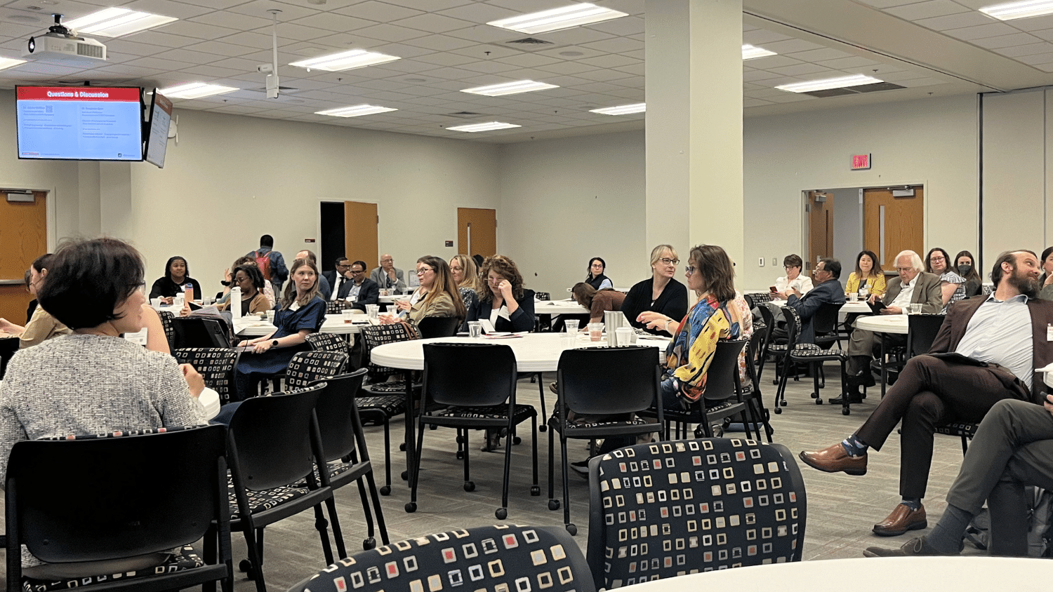 Professionals gathered around circular tables in a large conference room with grey walls. There are a few dozen adults sitting in black chairs facing a presenter towards the left of the frame.