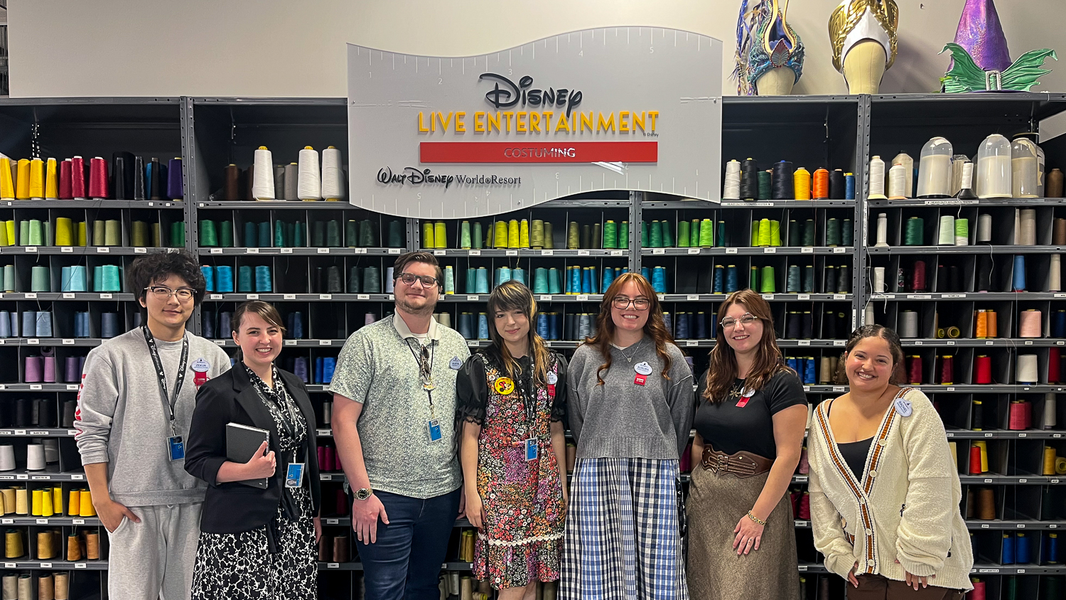 Jamie Piraino and six other people stand smiling in front of shelves filled with colorful thread spools under a sign that reads “Disney Live Entertainment Costuming Walt Disney World Resort."