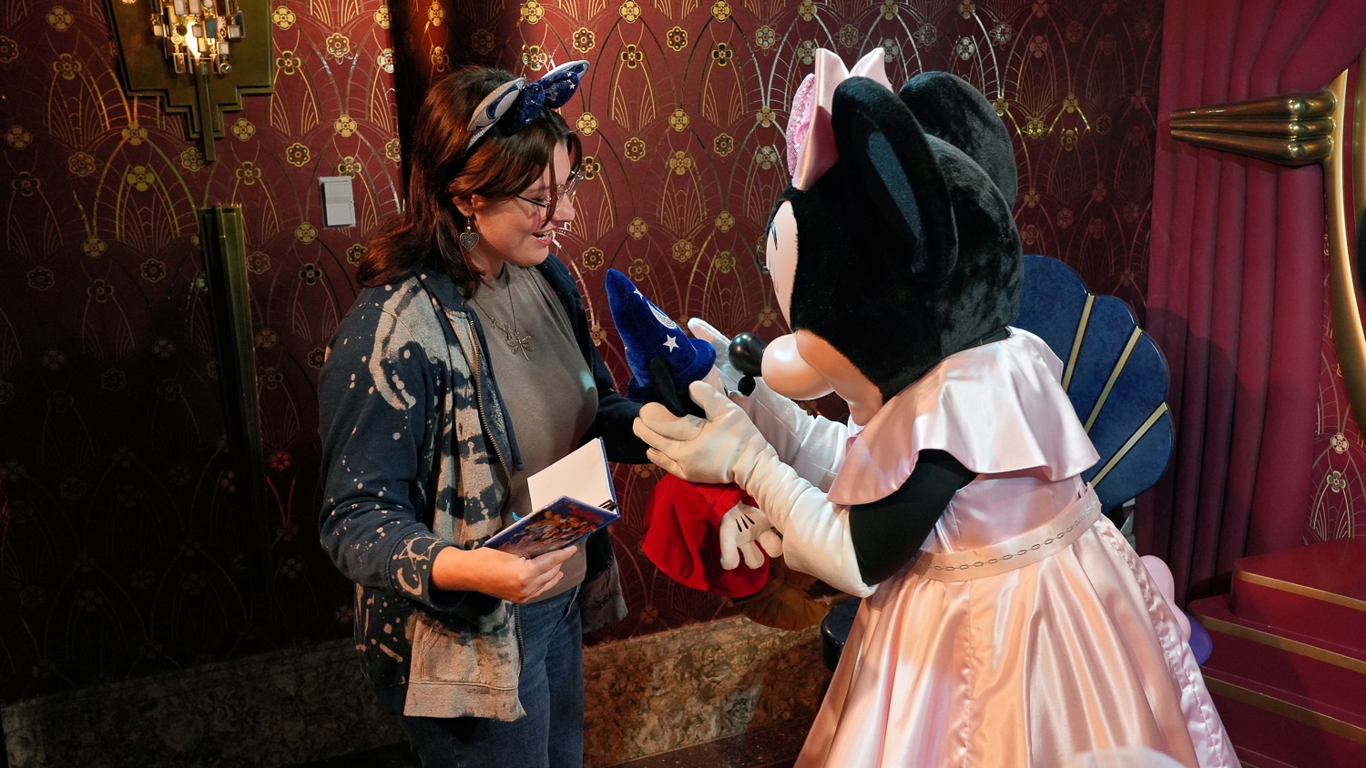 Jamie Piraino wears Minnie Mouse ears and glasses smiles as she meets Minnie Mouse, who is dressed in a white and pink gown, inside a themed, ornately decorated room. Piraino holds a book, a pen and a Mickey Mouse stuffed animal.