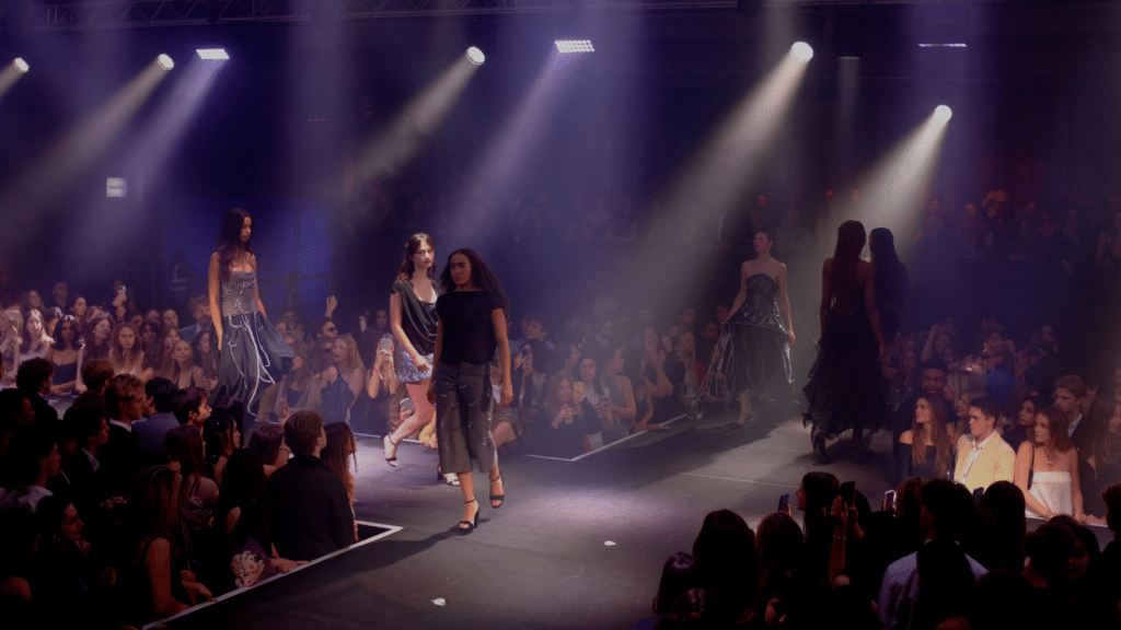 A wide view of models walkings on a runway bathed in purple and white light. The walkway is in the shape of an X and audience is packed into the space looking up at the models, who wear draped outfits with hardware features.