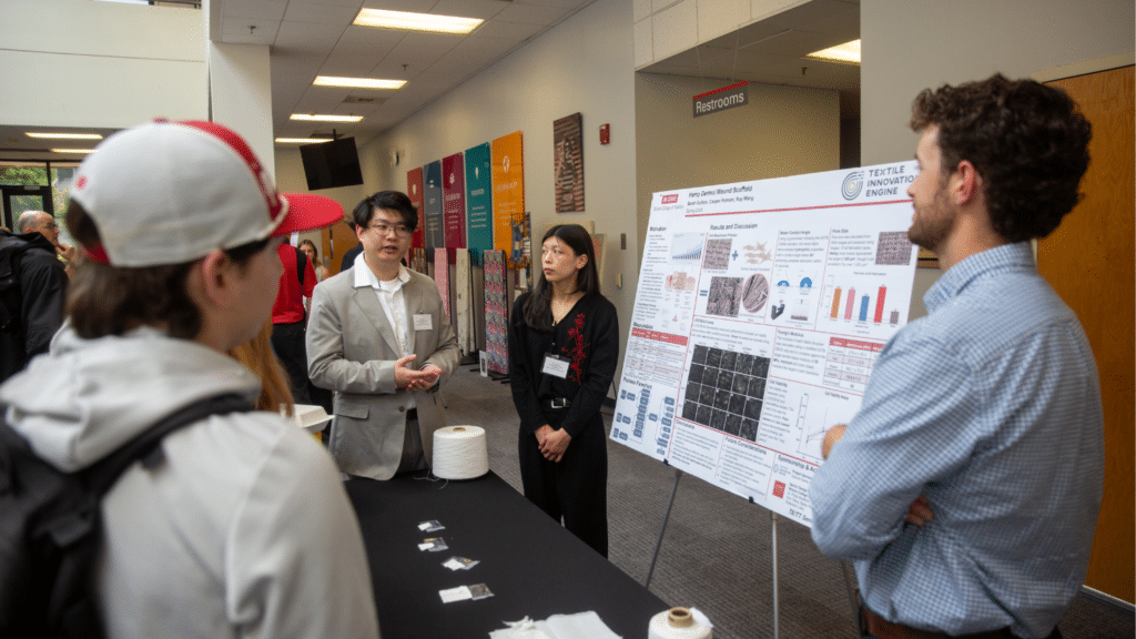 A student presents a large research poster to peers and judges, who stand around a table and face away from the camera. They are in a large lobby and surrounded by other attendees.