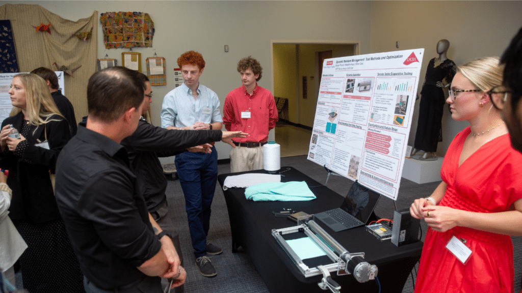 A student presents a large research poster to peers and judges, who stand around a table and face away from the camera. They are in a large lobby and surrounded by other attendees.