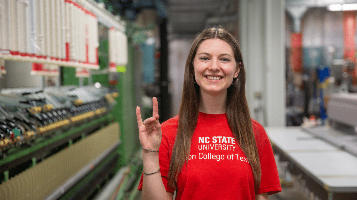 Kat Charrier holds up the "Go Pack" hand sign as she smiles at the camera and wears a bright red shirt. The complex machinery of the Spinning Lab is blurry behind her.