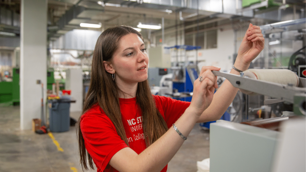 Kat Charrier wears a bright red shirt as she works with a machine in the Spinning Lab. She is looking at a spool of thread and delicately pulling the string towards her. The rest of the lab is blurry behind her.