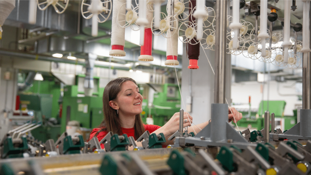 Kat Charrier smiles as she works with a machine in the Spinning Lab. There are mechanic components bordering the frame as the camera looks between them and focuses on Kat. The rest of the lab is blurry behind her.