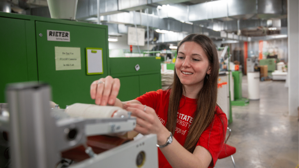 Kat Charrier wears a bright red shirt as she works with a machine in the Spinning Lab. The rest of the lab is blurry behind her.