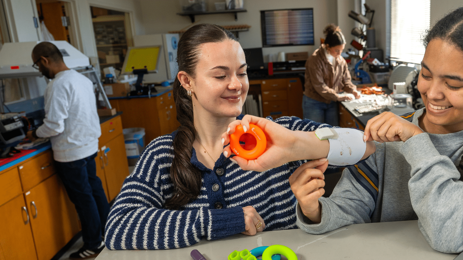 Sophia Thompson looks down at her forearm, which is being equipped with an armband with embedded technology. Her dark hair is in a slicked-back ponytail and she wears a navy and white striped sweater. To the right is another student fastening the armband, and a lab is laid out behind them with two other student in frame working on countertops.