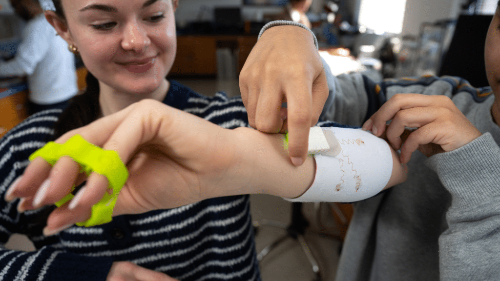 A close-up of Sophia Thompson looking down at her forearm, which is being equipped with an armband with embedded technology. Her dark hair is in a slicked-back ponytail and she wears a navy and white striped sweater. To the right is another student fastening the armband.