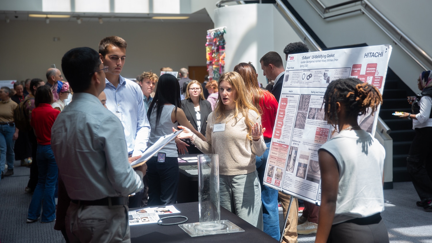 A student presents a large research poster to peers and judges, who stand around a table and face away from the camera. They are in a large lobby and surrounded by other attendees.