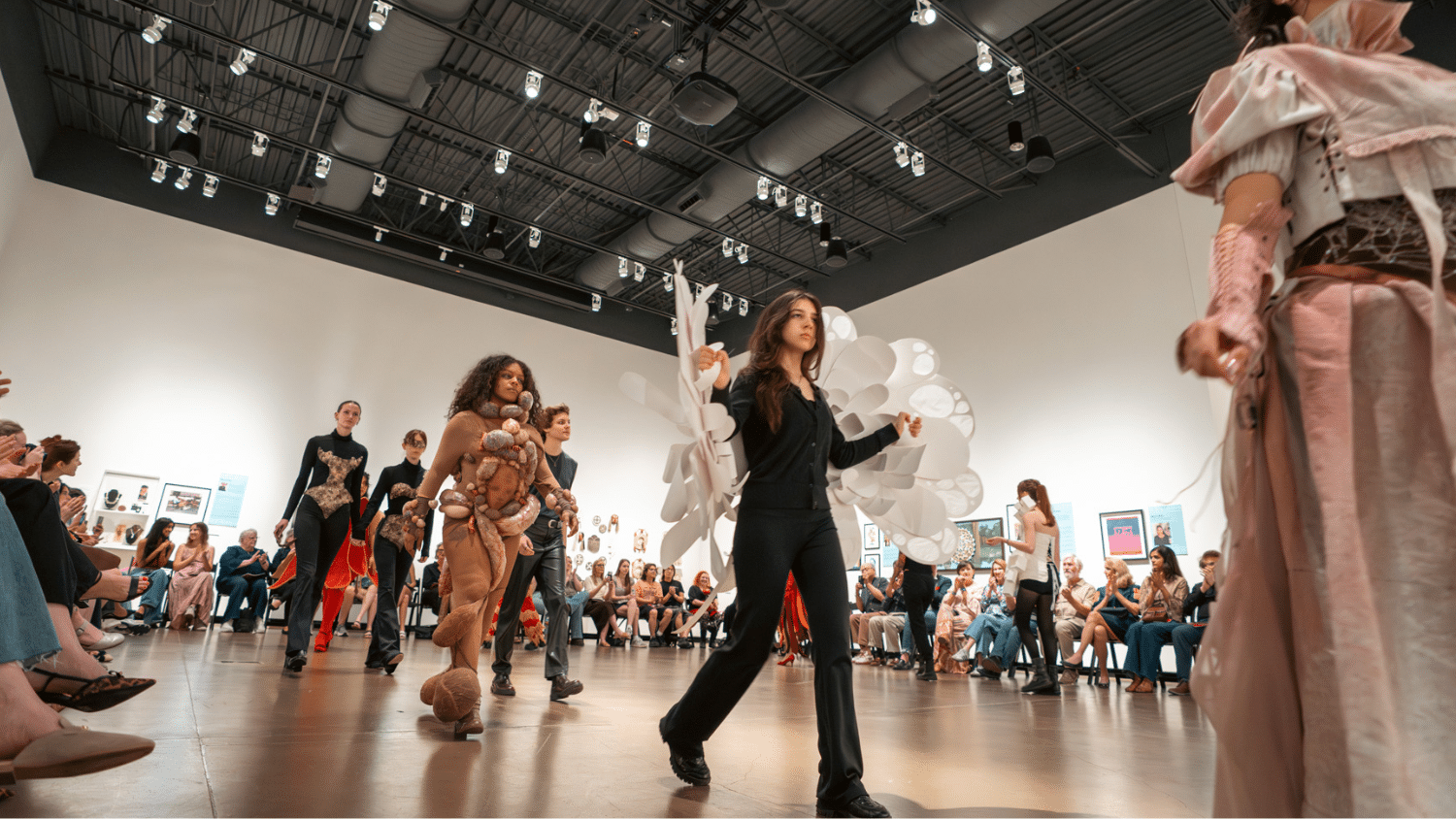 Students walk the final runway through an open room in the Gregg Museum of Art & Design. They are wearing elaborate costumes made of latex, paper, and textured fabrics. The room is bordered with sitting onlookers and walls of art.