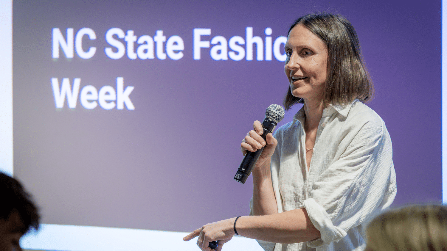 Kate Nartker stands in front of a projected screen that reads "NC State Fashion Week" with a microphone in hand. She has a shoulder-length hair and wears a white linen button up. She is speaking to an audience.