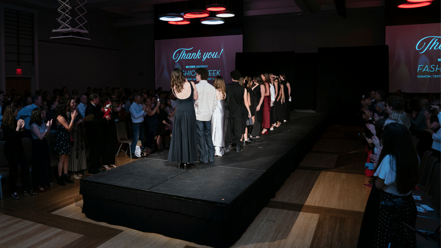 The designers from the Emerging Designers Runway Show line up on the runway in a dark room as the audience stands and claps for them.