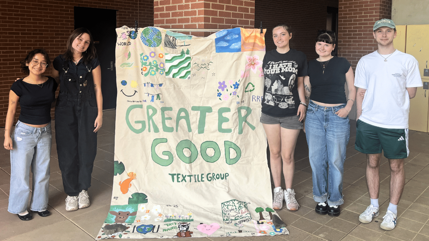 Five student pose with a large banner reading "greater Good Textile Group." The banner is painted with small designs of fish, water and trees.
