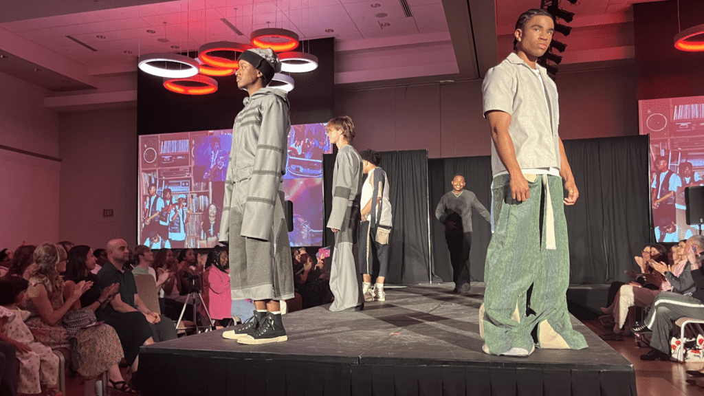 A students pose on the runway of the African American Textile Society's Fashion Exposé as the designer of their outfits walks down the center smiling.