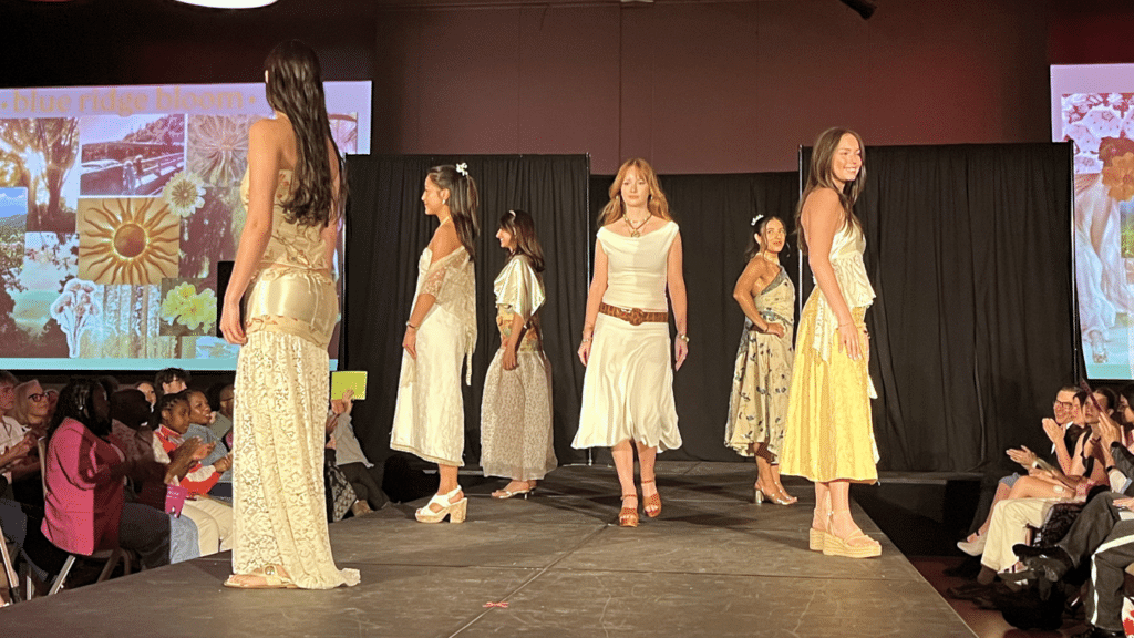 A students pose on the runway of the African American Textile Society's Fashion Exposé as the designer of their outfits walks down the center smiling.