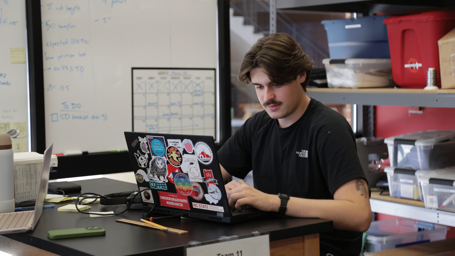 A student sits at a desk looking at his laptop, which is covered in stickers. He has short, brown hair and a mustache and wears a black tee shirt. In the background is shelves with bins and a whiteboard with a to-do list on it.
