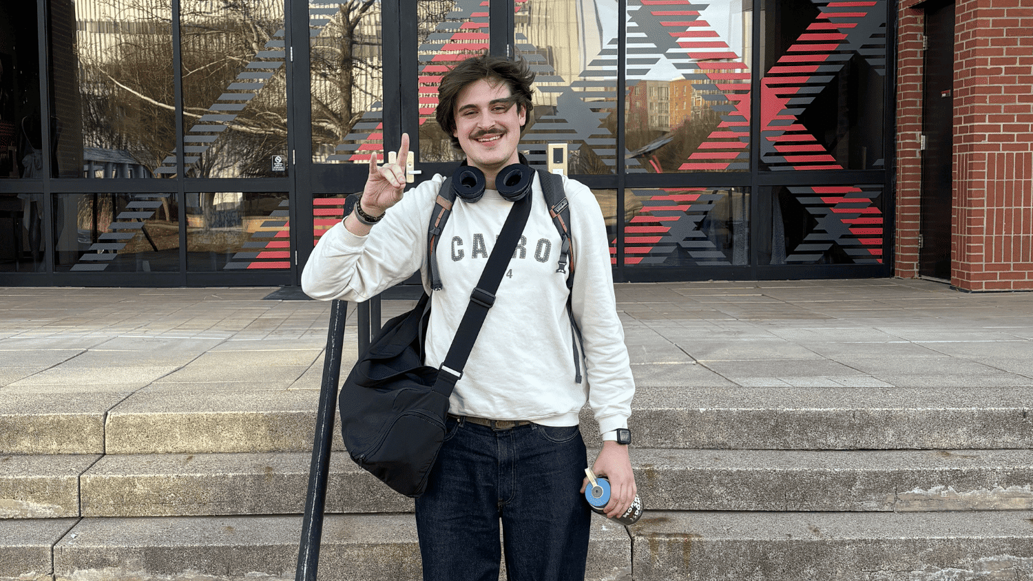 A student stands in front of the Textile Complex holding the Pack Pride hand gesture. He has a duffle bag over his shoulder, black over-ear headphones around his neck, and a backpack. He stands in the middle of a set of concrete stairs.