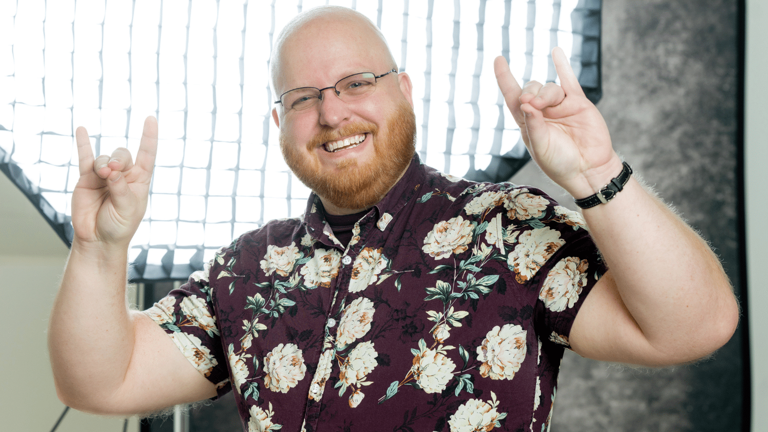 Andy Click smiles at the camera and poses with two "Go Pack" hand gestures. He wears a purple, floral, short sleeve button up and a black watch. He is bald and has a red beard and thin glasses. He stands in front of a large light that takes up most of the background.