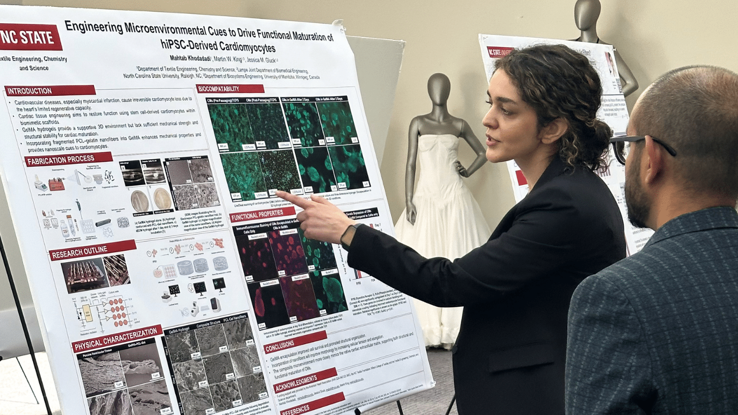 A graduate student presents a research poster to a audience member. She has long wavy brown hair tied back and wears a black blazer as she points to her poster. The bystander is in the foreground facing away from the camera. The poster is jammed with words and colorful images of microscopic fibers.