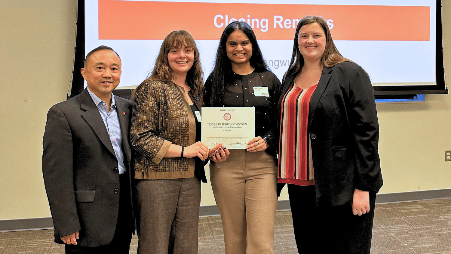 Two students stand between two officials from the Textiles Research Symposium. They both hold a certificate. Behind them is a blank wall with a projector screen reading "Closing Remarks."