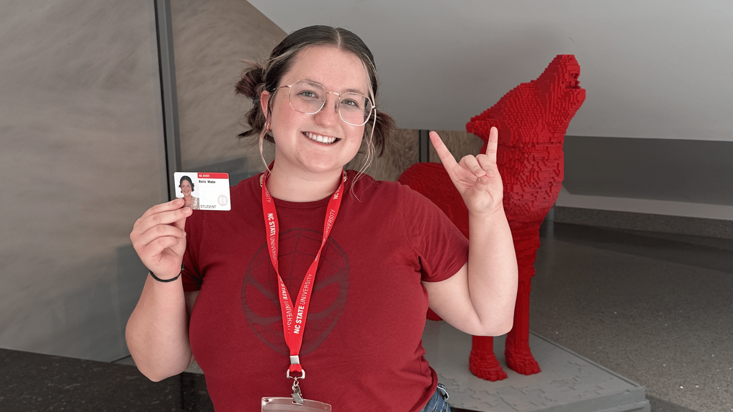Bella Mabe poses in a red t-shirt and her hair in two buns. She holds up her student ID and a wolfie hand signal. She stands in front of a red lego sculpture of a wolf.
