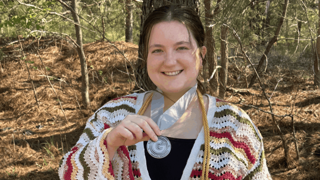 Bella Mabe poses in front of a wooded background and smiles at the camera. She holds up her graduation medal from the Gaston College. Her long brown hair is tied back and she wears a knitted cardigan.