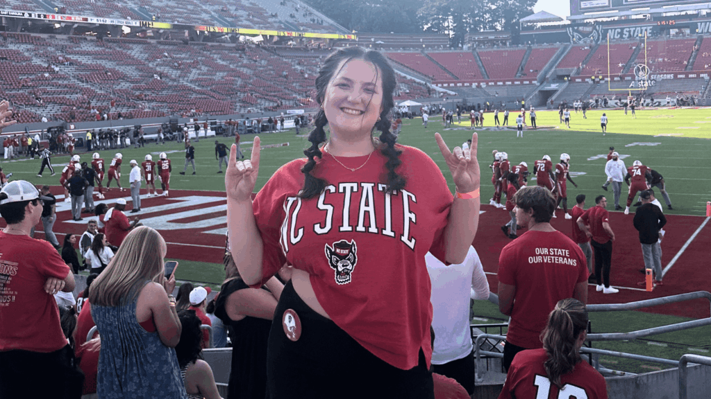 Bella Mabe holds up two wolfie hand signs and smiles for the camera. She wears an NC State branded t-shirt and stands in front of the NC State football field, the stadium full of players and people.