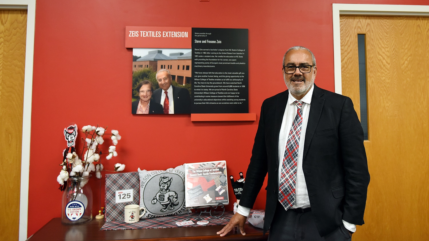 Andre West smiles for a picture in front of a display commemorating the Zeis Textile Extension. He wears a black suit with a red, black and white plaid tie. The table he leans on features Wilson College merch and a vase of cotton. The plaque on the red wall behind him features the photos of Steve and Frosene Zeis next to a block of text.