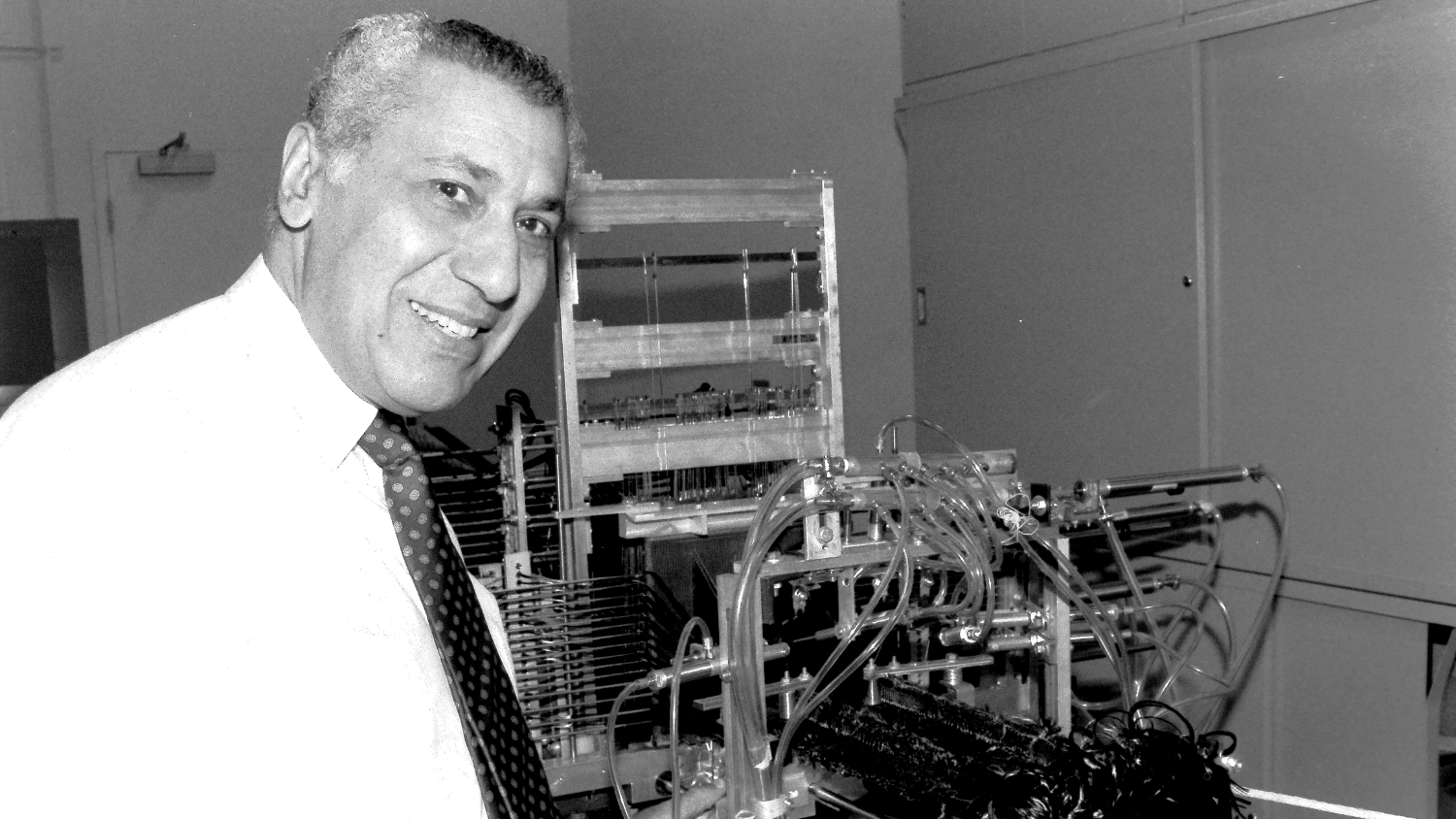 Mansour Mohamed smiles at the camera in front of weaving machinery. He wears a bright white shirt that takes up most of the left-hand corner of the black and white film photograph. The machine behind him has many tubes leading to a loom-like structure.